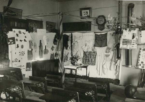 An autumn exhibit and fair has been set up in the Centerville School. A post supports streamers which extend to the walls, and examples of needlework and crafts are displayed. A pumpkin sits on the floor near a row of desks. There is clock on the wall and a chandelier with three oil lamps hangs from the ceiling. On the reverse of the photograph is written "School now closed, transporting to East Troy."