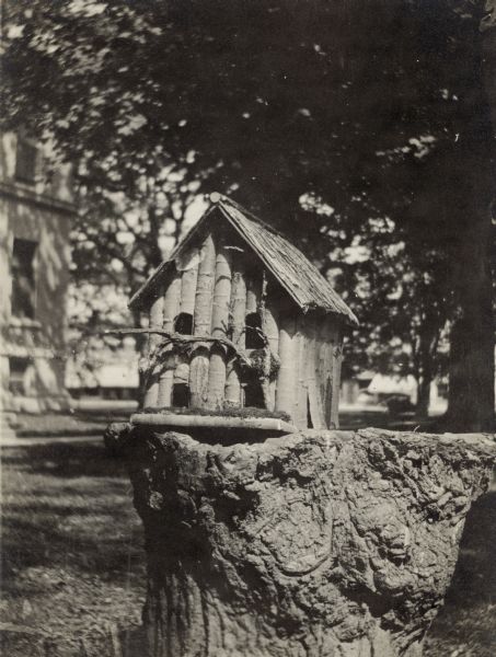 A rustic birdhouse with four separate entrances is displayed on a tree stump with gnarled bark. On the reverse of the photograph is written "Prize bird house at fair. One of the many patterns used. Bird houses [were] made in every school in connection with our 'School Ground Beautification and Bird Lure' projects from 1929 - 1932."