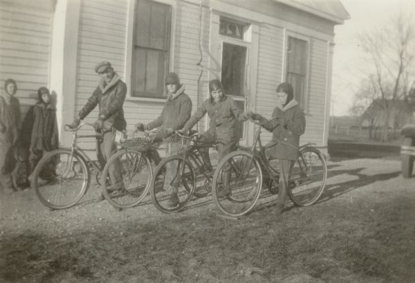 Four boys on bicycles pose at the South Heart Prairie School as two others look on. With bicycles are Charles Davis, Howard Holden, Ivan Bogie, and Raymond Buckholz. The boys are dressed for cold weather with hats and coats. It is noted on the reverse of the photograph that this is "One way pupils travel to school" and that the teacher at the time was Patricia Rotier.