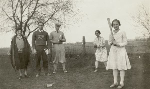 A girl poses with a bat as a second girl holds a ball. Two young men in sweaters, identified as students from Whitewater Teachers College (now the University of Wisconsin-Whitewater) observe. A woman with a cane stands next to them. The men acted as "director of games" at a rural school.