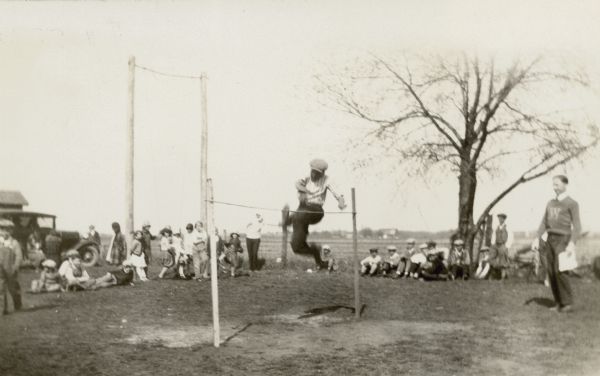 A boy is caught mid-air in his approach to the high jump. Many other children and adults watch. One man identifiable by the "W" on his sweater is a student at Whitewater Teachers College, now University of Wisconsin-Whitewater, and acted as "director of games" at a rural school.