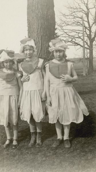 Faye Finch, Dorothy Tober and Doris Behling pose out of doors.  They are dressed in crepe paper costumes with petalled skirts, leaf bibs and caps with stems. Students at the Arrow Lodge School, they entertained at the Township Play Day.