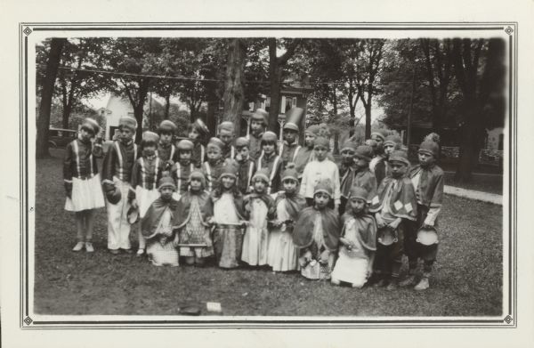 Members of the rhythm bands from the Woods and Fontana state graded schools pose outdoors holding their instruments. The children in the front row and on the right wear uniforms with capes and caps with tassels; the others have matching jackets and flat caps. They are photographed on the lawn of a large brick farmhouse. A barn and an automobile are in the background. The bands took part in the County Music Festival.
