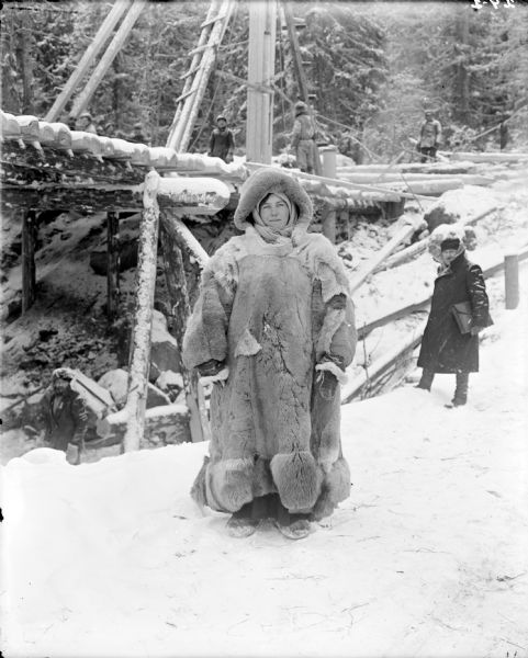Portrait of of a Russian woman wearing heavy furs. She is standing near the construction of a wooden bridge. In the background are men standing near and on the bridge.