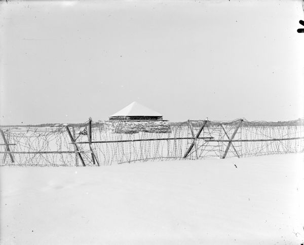 View across snow and through barbed wire fence towards a timber constructed blockhouse. More buildings and a forested area are in the far background.