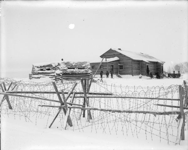Blockhouse, Standard Hut, and Kitchen Building View over barbed wire fence towards men standing outside a blockhouse and barracks. Immediately outside of the barracks is a horse-drawn mobile kitchen and harnessed horse.