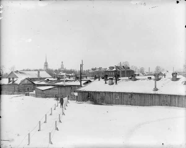 Quarters of 310th U.S. Army Engineers Elevated view of the quarters in Solombola, Russia for Company C of the 310th United States Army Engineer Corps. A uniformed soldier stands in the snow outdoors within a fenced pathway leading to the quarters. In the background are buildings with towers that rise above the trees.