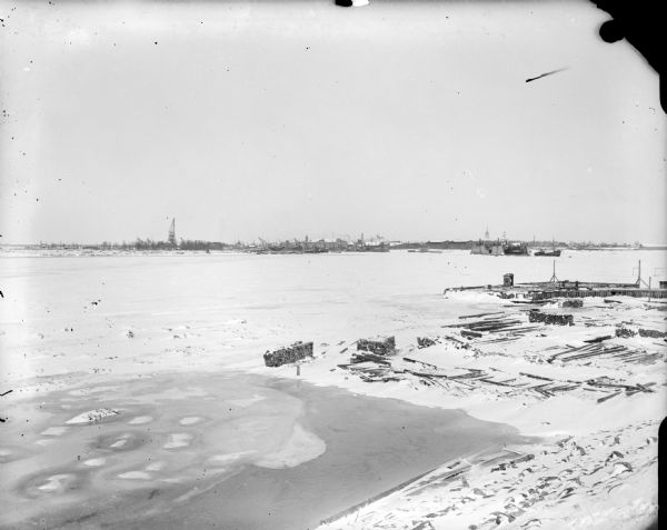 View of Solombola from Archangel [Archangelsk], Russia. In the distance are series of cranes. In the foreground on the ice are stacks of logs near a pier made of pilings. Just beyond the pier on the opposite shoreline there appears to be a vessel in dry dock, and another vessel next to it.
