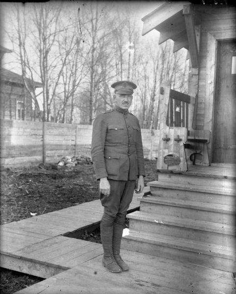 Outdoor portrait of Captain Armstrong of Company C from the 310th United States Army Engineer Corps, standing on a board sidewalk in his uniform. He is standing in front of a wood building with a small front porch with a bench which has the United States Army Engineers emblem carved out of the back. In the background across the yard is a wooden fence and behind it another building.