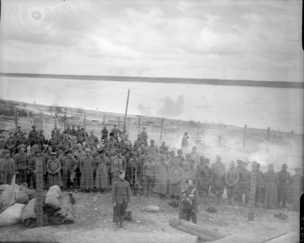Elevated view of a group of Bolshevik prisoners being kept near a shoreline in a barbed wire enclosure. They are being guarded by a group of armed American, British, and Scottish soldiers.