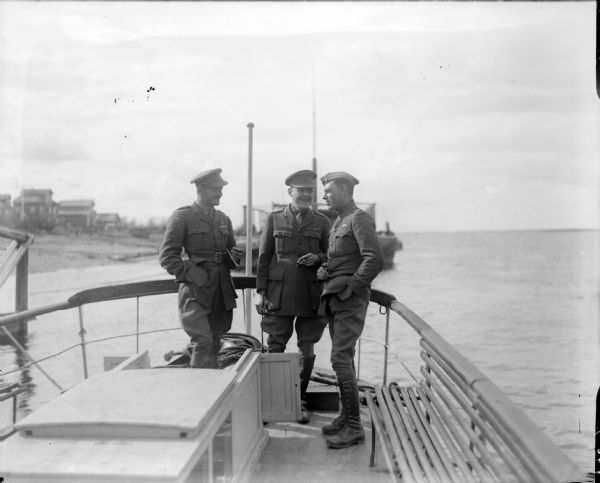 Major McArdle with two British officers in uniform on board the launch "Wisconsin." All three men are smoking cigarettes. Another ship and a shoreline can be seen in the background.