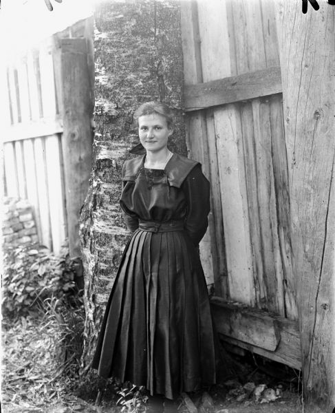 Portrait of a woman wearing a long dark dress standing in front of a birch tree and a wooden fence.
