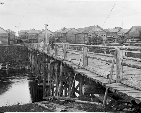 Soldiers Sitting on Bridge View from shoreline a group of British soldiers sitting on the railing of a wooden bridge that crosses a river. In the background on the opposite shore is a village with many wooden buildings. A Russian woman is standing on the bridge in the background, and a group of people are walking along the path towards the bridge.