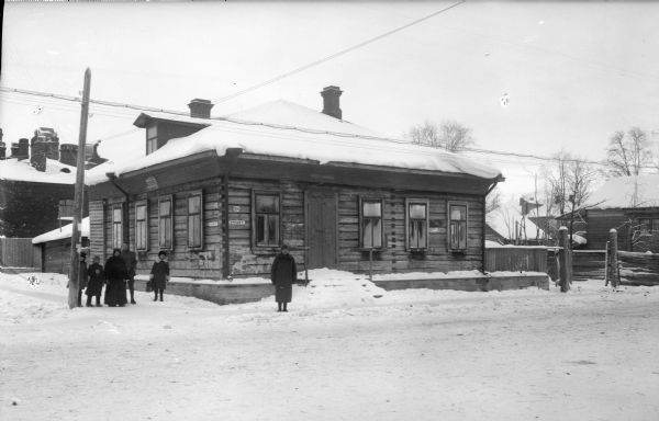 View from snowy road of the exterior of the sleeping quarters and mess of the United States Army Engineer Corps. There is a Russian family standing alongside the building. A soldier stands amongst them. There are Russian signs posted on the building.