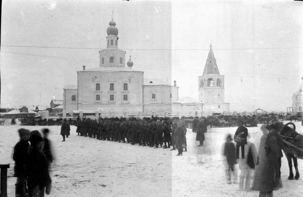 A large group of Russian soldiers are assembled and appear to be drilling within the center of a Russian town. Civilians stand off to the side observing the soldiers. In the background is a large number of horses and sleighs near a stone wall with a railing. Behind the stone wall is what appears to be a church building with onion domes, and a bell tower with arches.