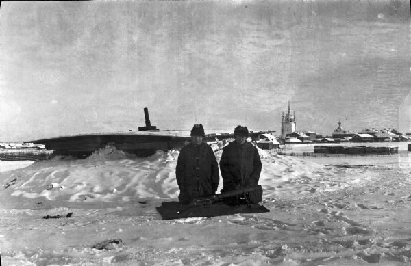 Two soldiers wearing heavy winter clothes are kneeling behind a Lewis machine gun displayed on a blanket. Immediately behind the two soldiers is what appears to be a fortification hidden with snow, but with the roof and chimney pipe showing. In the far distance is a town with a number of buildings. The tallest building appears to be a church, and has arches, towers, and small onion domes.