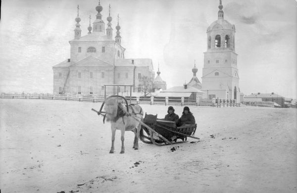 Two officers wearing heavy winter clothes are seated in a horse-drawn wooden sleigh on a snowy path. Just behind them is a building that appears to be a Russian church with small onion domes, and a fence, bell tower, and other buildings.