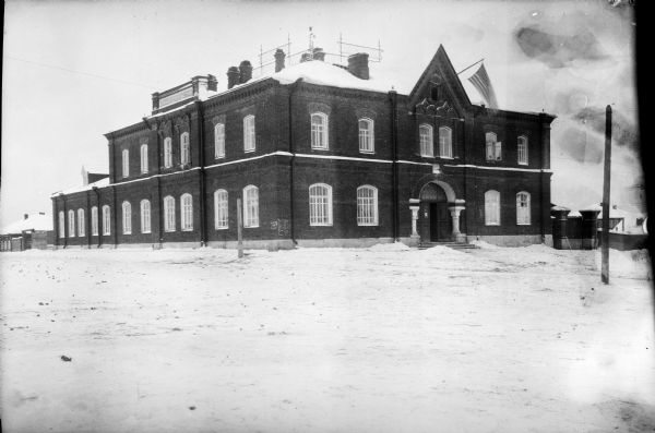 U.S. Army Headquarters Exterior view across snow of the headquarters building of the United States Army during the Allied Intervention in Northern Russia. An American flag is flying from an open window near the roof of the two-story brick building, which has an arch and two columns framing the entrance.