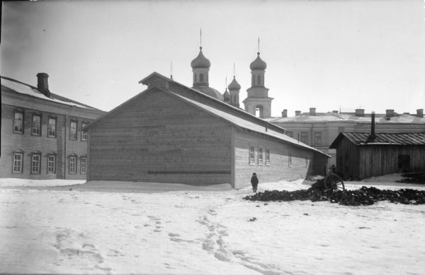 View across snow towards a child standing near the United States Army machine gunnery school, which is surrounded by other buildings. The towers and onions domes of a Russian church can be seen behind the building in the center. Next to the gunnery school building is a wrecked caisson and a shed.