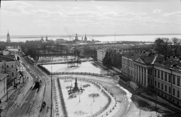 Elevated view of Troitsky prospect in Archangel [Archangelsk], Russia, looking south, showing homes, churches, state buildings, as well as the statue of Peter the Great. In the distance are cranes from the city's port area. There is also a streetcar passing pedestrians walking on the sides of the road.