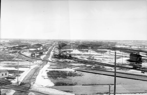 Elevated view of a series of three rows of large buildings, possibly used to store supplies. There are railroad tracks alongside each of the buildings. Snow and ice cover the ground.