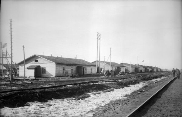 A group of barracks near railroad tracks with snow in the ditch. On the far right three soldiers are walking along the railroad tracks, and other men are near the barracks.