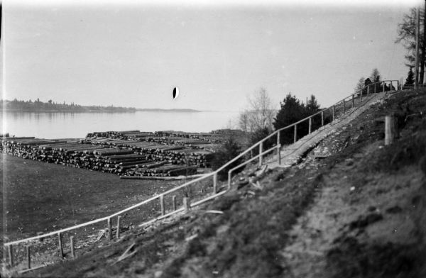 Looking toward the Pinega River, showing the stairs and path to its shoreline, as well as a stacks of logs piled up along the shore.