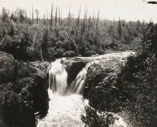 Elevated view of Copper Falls on the Bad River. Four people, one man and three women, are posing on rocks at the top of the falls. Surrounding burned-over forest area.