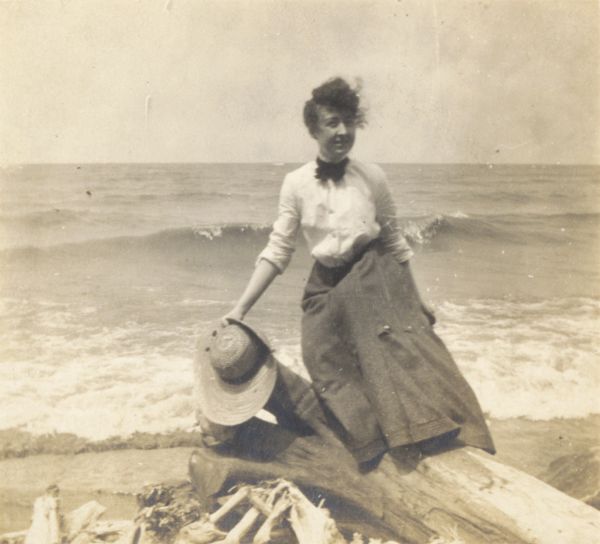Young woman on a driftwood stump, standing at the shoreline with what is probably Lake Michigan behind her. She is wearing a long skirt and a blouse, with a dark neck bow.

