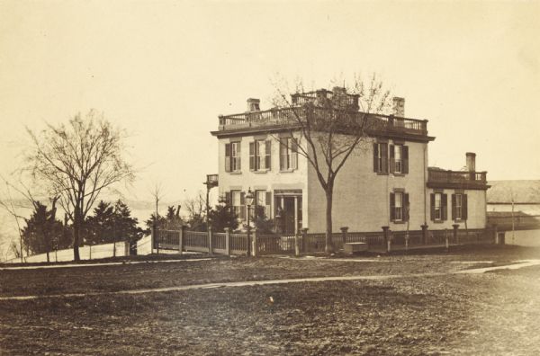 View across lawn of the residence of Lucius Fairchild, in Madison. A woman is standing outdoors near the entrance. A fence surrounds the yard of the house, which has a widow's walk on the roof. There is a lake and far shoreline in the background.