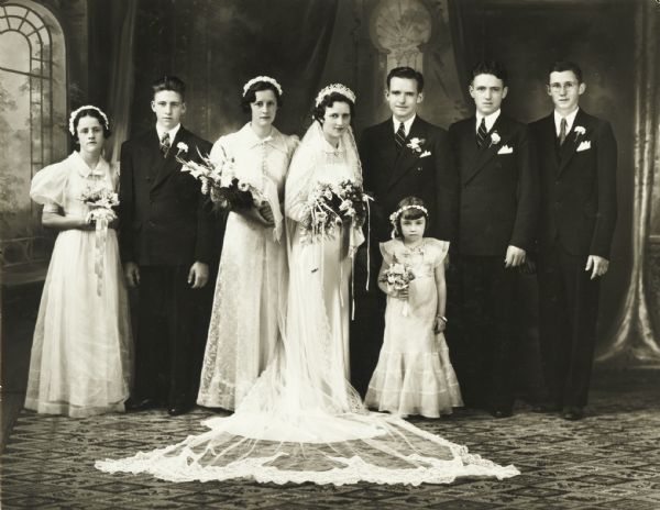 Studio group portrait of wedding party all standing while posing in front of a painted background. Three women and a young girl are wearing long white dresses and are holding flowers. Four men wear suits with boutonnieres in their lapels.