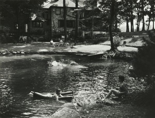 Suburban upper-class residence in wooded area with rock-lined swimming pool and swimming children. Two women sit in lawn chairs on the lawn near the house in the background.