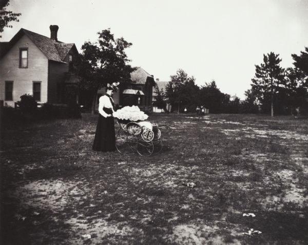 Young woman in residential area, standing in a yard pushing a baby carriage with a ruffled top. On the left are houses, and in the far background a group of people are standing near trees.