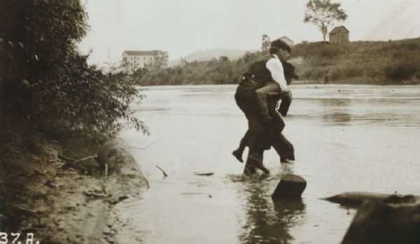 Man carrying another man on his back near the shoreline of a river. In the background on the far shoreline are buildings.