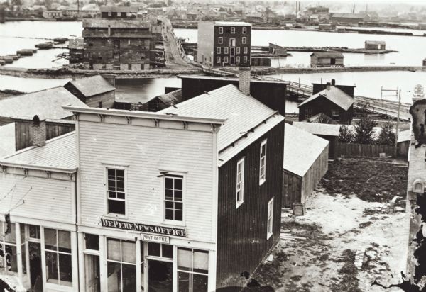 Elevated view over De Pere. In the foreground is the De Pere News Office and Post Office building. Behind it people are walking on the bridge over the Fox River. On the far shoreline are buildings and dwellings. Buildings and sheds sit on pilings on both sides of the bridge.