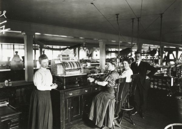 Three women and one man pose at a cashier's counter in the interior of a dry-goods store, presumably in Wisconsin. Hammocks hang from the ceiling in the background.