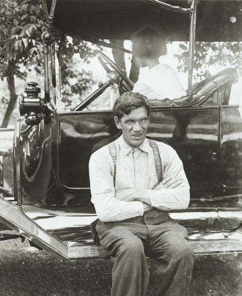 View of a man, who is apparently blind in one eye, sitting on the running board of a Model T Ford touring automobile outdoors. A young girl is sitting in the driver's seat.