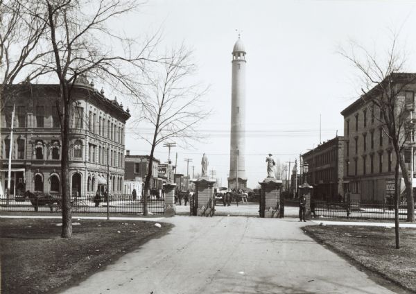 The Madison water tower and farmers' market as seen through the stone gateway at the East Washington Avenue entrance to the Capitol Park. The building on the left became the American Exchange Bank in 1922 when the First National Bank moved into their new building across the street.