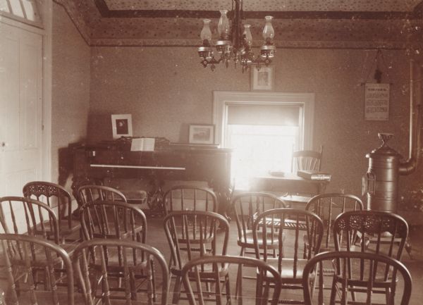 "Society Room" at Downer College. The view from the back shows chairs lined up in rows, a chandelier hanging from the ceiling, and a piano against the wall in the corner on the left, a desk and chair in front of a window, and a wood burning stove in the right corner.