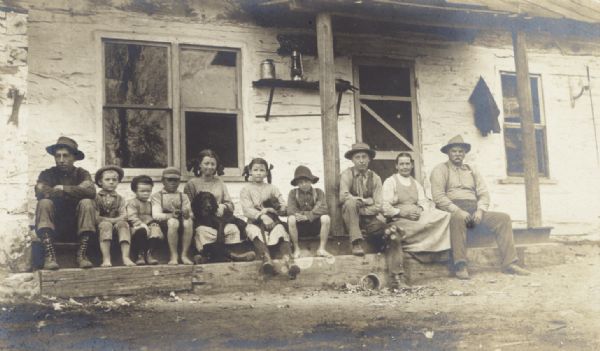Hansens family sitting in a row along the front of a porch with Mr. and Mrs. Phelps. One of the girls has a dog in her lap, and another girl has a puppy in her lap.