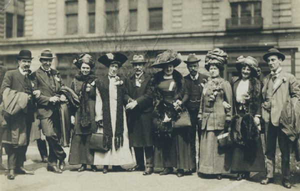 Group of well-dressed men and women wearing hats posing outdoors standing in a line, probably on a street in Milwaukee. Possibly a group of convention delegates, since they all are wearing badges. The women are all wearing long dresses and coats, and some have fur stoles and fur hand muffs.