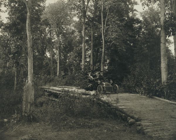 Dr. W.T. Rinehart, a prominent Ashland physician and surgeon, with Mrs. Rinehart and a sister, in an automobile crossing a small log bridge, probably over Deer Creek, south of Ashland. A dog stands on the bridge in front of the automobile.