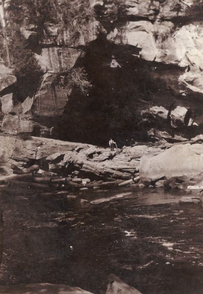 Ontonagon River View from rocky shoreline across river towards a man standing on rocks and holding a stick in front of a rock face on the opposite shoreline. There are large logs piled in the water below the man.