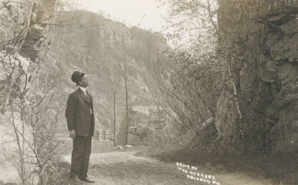 A man wearing a suit and hat is standing in a roadway through "the narrows." In the background is a steep ridge. Also in the background are stacks of wood and an electric power pole.