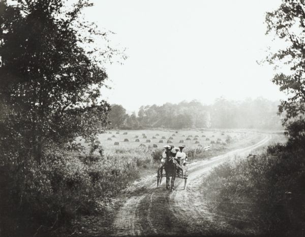 View down tree-lined country road near Wausau, taken by Dr. Joseph Smith, talented amateur photographer. It depicts women (probably members of his family) driving a horse and buggy. In the background is a field of grain. The picture captures the spirit of the pleasure drive movement of the 19th century, but it was by no means typical of the road conditions faced by Wisconsin travelers.