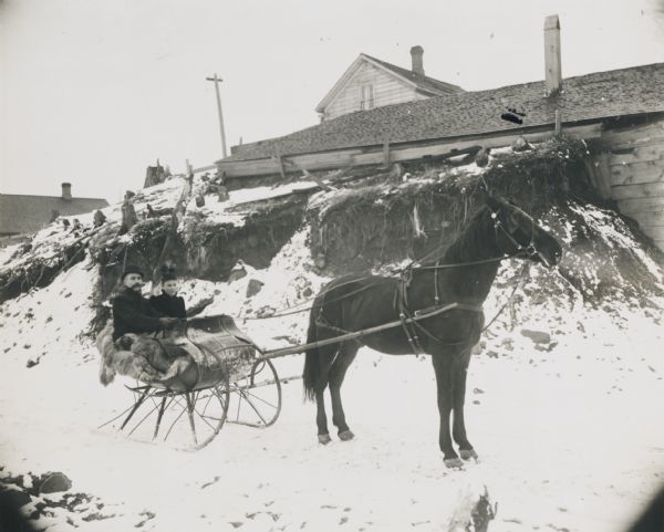 Winter scene with a man and woman driving in a horse-drawn "cutter" in the snow. They have a fur blanket over their laps. Behind them is a building, perhaps a barn, built into the side of an embankment.