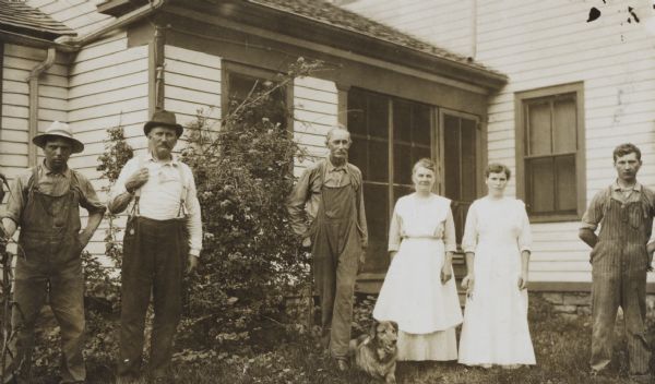 Mr. Gilbert's family standing in front of a farmhouse. The man on the left is holding what appears to be a cornstalk. Three other men, two women, and a dog are posing in the yard.