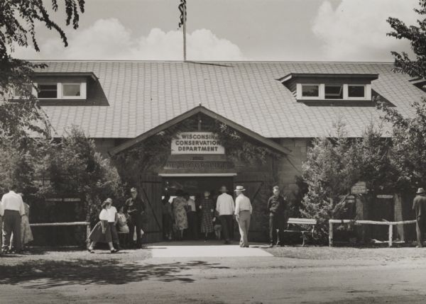 Entrance to a building housing "Outdoor Life Exhibits" by the Wisconsin Conservation Department. A number of people are in front of the entrance, and more are entering through the open doorway. Two men in uniform stand by benches flanking the entrance. Fences on both sides of the building enclose animal exhibits, and the one on the right is marked "Black Raccoon."