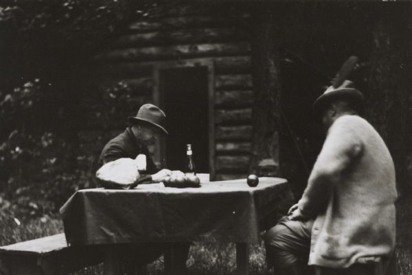 Two men are sitting on benches at a table in front of a fishing camp log cabin in northern Wisconsin. Both men are wearing hats, and the man on the right has a large feather on his hat.