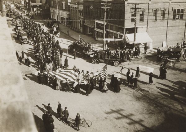 Elevated view from building of informal Armistice Day street parade at an intersection of the downtown area. They are celebrating the conclusion of the first World War. A group of women are walking while holding onto the sides of a large flag. Other marchers carry U.S. flags. Flatbed trucks going in the other direction carry groups of people who are standing and holding onto the sides. Pedestrians watch from the street and the sidewalks.